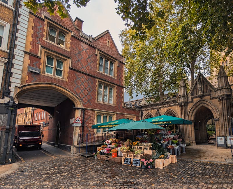 A vibrant flower stall situated on a cobblestone street at the base of a red-brick building with an archway, featuring a large green umbrella displaying the website URL 'earlsfieldflowers.co.uk.' The