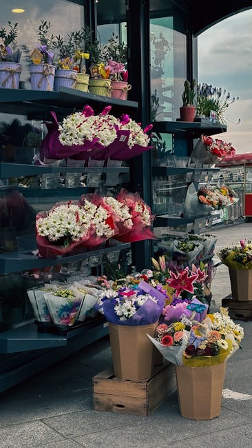 A display of various floral bouquets and potted plants arranged on black metal shelves inside a glass-walled shop, with a focus on fresh flowers including white chrysanthemums, pink carnations, purple
