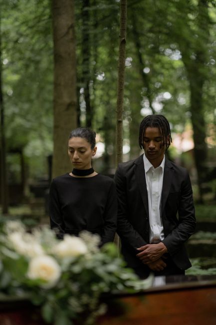 A somber scene featuring two individuals dressed in black, standing with heads bowed in a solemn moment, possibly at a funeral or memorial service. In the foreground, a floral arrangement of white ros