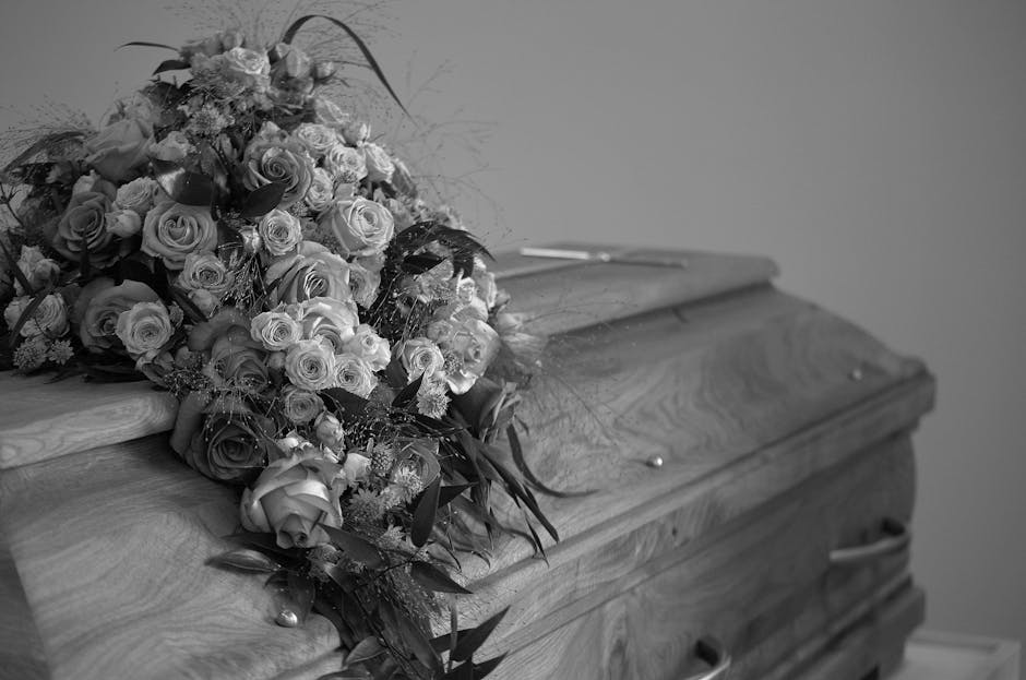 A large, lush bouquet arranged with various roses and other flowers, featuring a mix of open blooms and buds, placed on a wooden casket. The flowers include roses in different sizes, with some petals