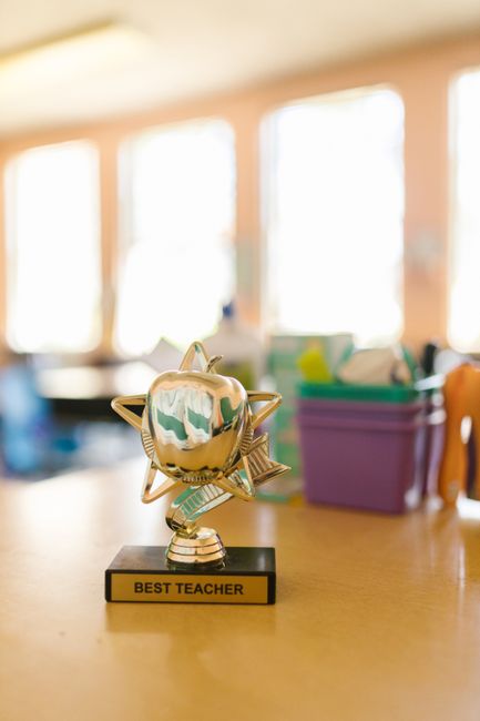 A shiny gold trophy with a metallic apple at its center, surrounded by star-shaped accents, placed on a black base with a yellow plaque engraved with 'Best Teacher'. The background features blurred cl