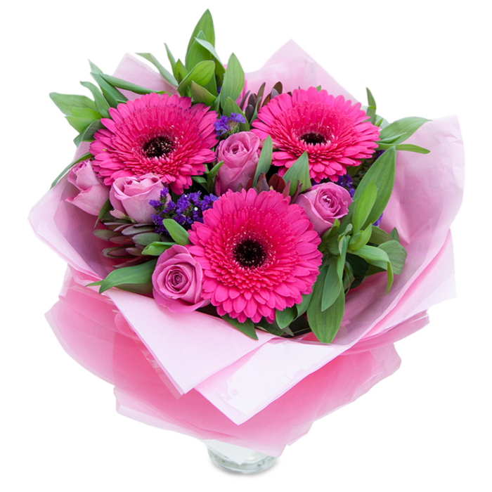 I picture three vivid fuchsia Gerbera daisies taking centre stage, each blossom opening wide with layered, radiating petals encircling a dark brown to black eye that anchors the composition. Around those confident blooms, lavender-pink roses unfurl in a slightly muted, dusty-rose tone, their inner petals still tucked in that fresh-bud way that promises a gentle, velvety touch. Little clusters of purple-blue statice are tucked throughout like confetti, their papery texture and cool hue offering a charming contrast to the softer roses and the saturated Gerberas. The greenery-broad salal-like leaves and streamlined eucalyptus-sits lush and supportive behind the flowers, the leaves' cool green tones adding depth and a hint of dewy freshness. The bouquet is presented with care: an inner matte pale-pink tissue wraps the stems, then a translucent fuchsia-pink cellophane, slightly crinkled, is folded and arranged to give the whole piece a soft shimmer and cohesive pink theme. The visible rim of a clear glass vase at the base tells you this is meant for immediate display, stems trimmed and arranged for balance and longevity. In the calm morning light the arrangement feels playful yet graceful, as if it could brighten a bedside table or greet someone stepping off the train at nearby Earlsfield Station. The scent is gentle-roses offering a delicate perfume, gerberas a faint green note-creating a bouquet that communicates affection, celebration, and thoughtful care for birthdays, anniversaries, or 'thinking of you' moments.